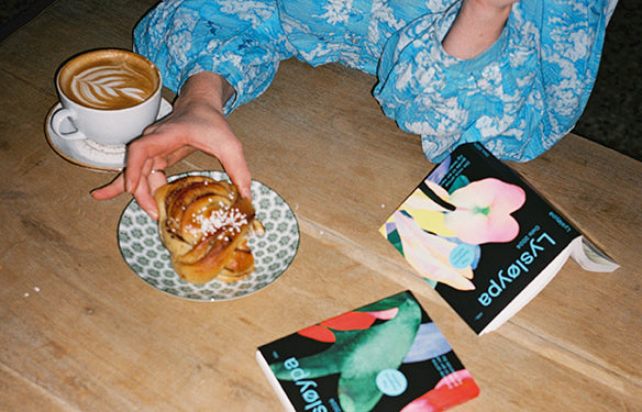 Kvinne sitter på kafé med Lysløypa bøker på bordet, drikker kaffe og spiser bolle